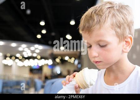 Ritratto di un piccolo ragazzo affamato divertente mangiare gelato freddo delizioso in una tazza di waffle seduti in un caffè, sfondo scuro, bokeh. Un bambino gode di cre di ghiaccio Foto Stock