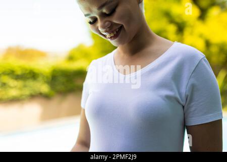 Felice donna biraciale che indossa una maglietta bianca sopra la piscina in giardino Foto Stock