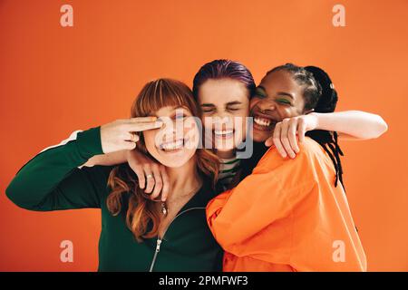 Tre amici femminili multiculturali sorridenti e abbracciati in uno studio. Gruppo di giovani donne vibranti che si godono mentre si levano in piedi di nuovo Foto Stock