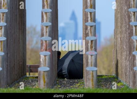 Dreieich, Germania. 13th Apr, 2023. Un uomo siede sotto un ombrello tra i tronchi della piramide della canna e guarda lo skyline di Francoforte sul meno. La Piramide dei Polacchi fa parte del 'Parco Regionale RhineMain'. Credit: Andreas Arnold/dpa/Alamy Live News Foto Stock