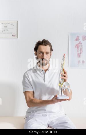 fisioterapista bearded che tiene il modello della colonna vertebrale e che guarda la fotocamera in sala di consulenza, immagine di scorta Foto Stock