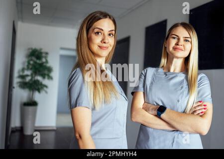 Belle donne sorridenti medici in uniforme medica stand vicino alla finestra in clinica. Una giovane infermiera sta parlando. Giorno di sole. Illuminazione interna. Spazio di copia. Foto Stock