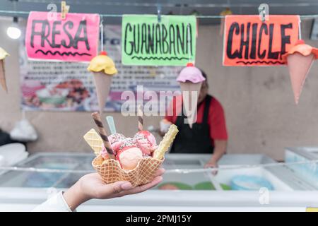 Una mano tiene un tradizionale gelato messicano decorato di fronte ad una bancarella di mercato di strada con segni di sapore Foto Stock