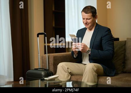 Il maschio con una valigia da viaggio siede in una poltrona in una camera d'albergo Foto Stock