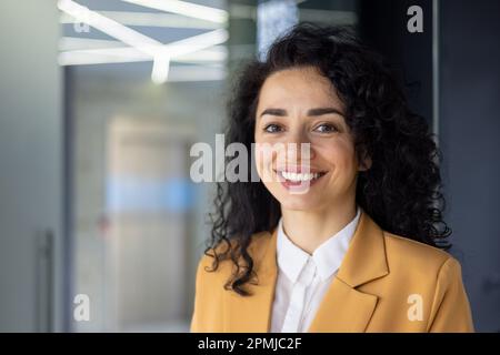 Primo piano ritratto di una donna adulto maturo business, capo sorridente e guardando la macchina fotografica, soddisfatto donna ispanica con capelli ricci in ufficio sul posto di lavoro. Foto Stock