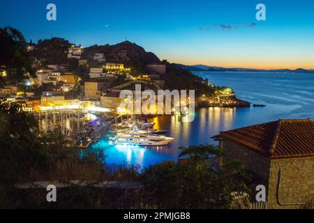 Tramonto su Hydra, una delle isole greche, e yacht nel porto e nel continente della Grecia in lontananza. Città illuminata Hydra su ripido pendio in blu h Foto Stock