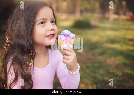 Ragazza carina bionda con capelli ricci mangiare un bel gelato nella foresta in una bella giornata estiva, il concetto di giorno dei bambini, ragazza felice godendo di una giornata calda. Foto Stock