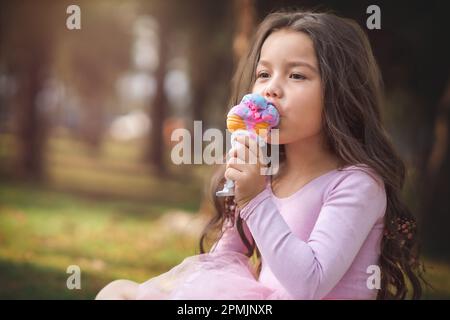 Ragazza carina bionda con capelli ricci mangiare un bel gelato nella foresta in una bella giornata estiva, il concetto di giorno dei bambini, ragazza felice godendo di una giornata calda. Foto Stock