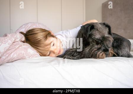 Carino bambino biondo, ragazzo toddler, sdraiato sul letto con il suo cane da compagnia a casa Foto Stock