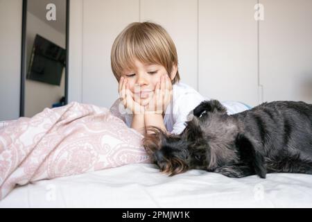 Carino bambino biondo, ragazzo toddler, sdraiato sul letto con il suo cane da compagnia a casa Foto Stock