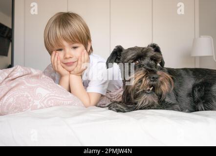 Carino bambino biondo, ragazzo toddler, sdraiato sul letto con il suo cane da compagnia a casa Foto Stock