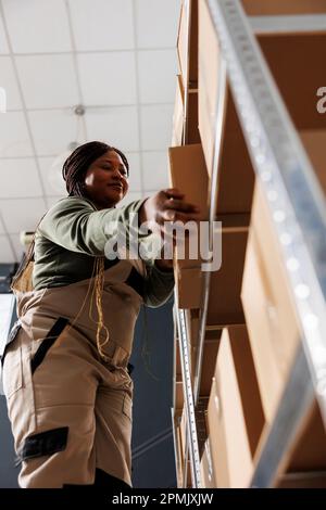 Dipendente del magazzino che preleva la scatola di cartone dallo scaffale metallico, preparando gli ordini dei clienti nel magazzino. Addetto al magazzino che indossa tuta industriale che lavora alla consegna delle merci Foto Stock