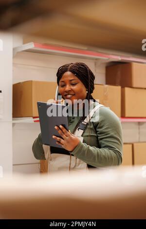 Dipendente afro-americano che discute dell'inventario dei prodotti con il manager remoto utilizzando un telefono fisso, controllando l'ordine online del cliente sul tablet. Supervisore che lavora in magazzino Foto Stock