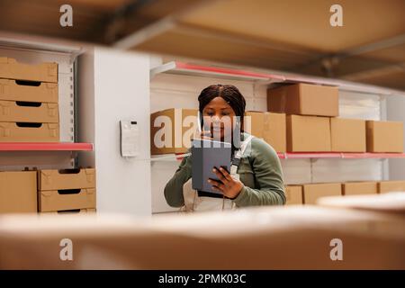 Un lavoratore afroamericano che parla al telefono fisso con un manager remoto, discutendo l'inventario dei prodotti in magazzino. Dipendente che indossa un abbigliamento industriale per il controllo generale degli ordini online su un computer tablet Foto Stock