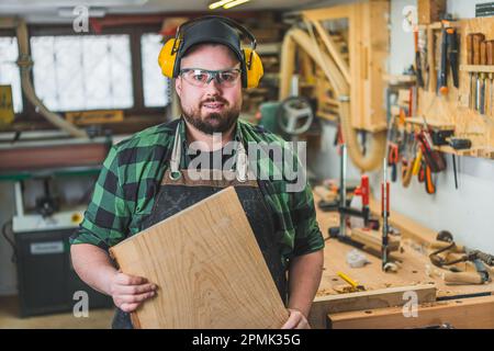 Concetto di lavoratore del collare blu. Carpentiere maschio focalizzato positivo in occhiali protettivi e cuffie che tengono la tavola di legno di fronte a lui. Foto di alta qualità Foto Stock