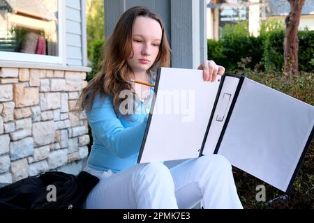 ragazza adolescente sfogliando attraverso il libro del notebook che tiene la matita imparare a sedersi sulla strada vicino alla scuola desiderio di studiare eccellente studente corretto bambino blusa blu capelli sciolti 12-15 anni spazio per il testo dell'annuncio Foto Stock