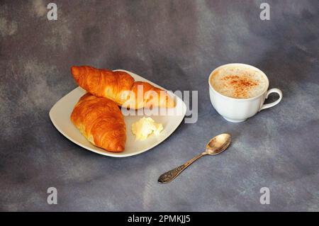 Una tazza di cappuccino caldo con cannella e un piatto con due croissant freschi con un pezzo di burro su fondo grigio. Primo piano. Foto Stock