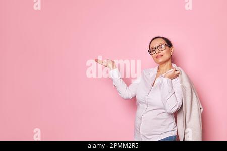 Donna incinta sicura in occhiali alla moda, tenendo spazio copia immaginaria sulla sua mano palmo in su, su sfondo rosa isolato Foto Stock
