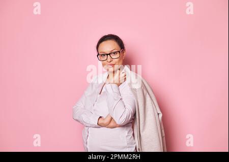 Fiducioso ritratto di una bella donna di mezza età in abiti alla moda e occhiali, guardando la macchina fotografica, isolato su rosa Foto Stock