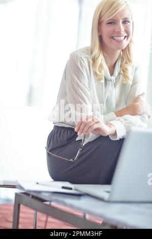 Buon lavoro qui. Ritratto di una donna sorridente che è seduta sulla scrivania di fronte al laptop. Foto Stock