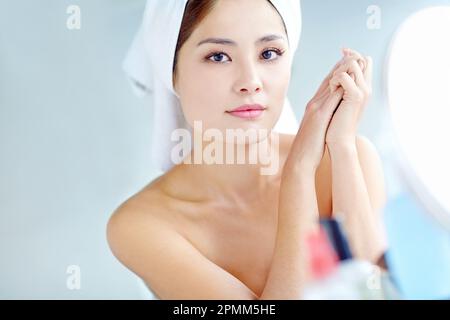 Idratante per le mani. Una bella giovane donna asiatica sorridente mentre si siede davanti a uno specchio con un asciugamano sulla testa. Foto Stock
