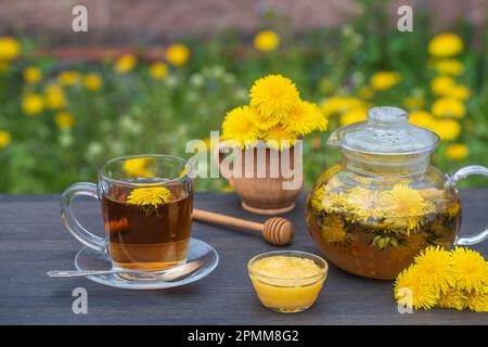 Delizioso tè alle erbe fatto di fiori di dente di leone fresco con miele su tavolo di legno in giardino, primo piano. Tè caldo dente di leone in teiera di vetro e tazza Foto Stock