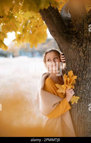 La bionda si trova vicino all'albero autunnale. Donna premurosa guarda avanti, vestita con un abito giallo. Contenuto autunnale. Foto Stock