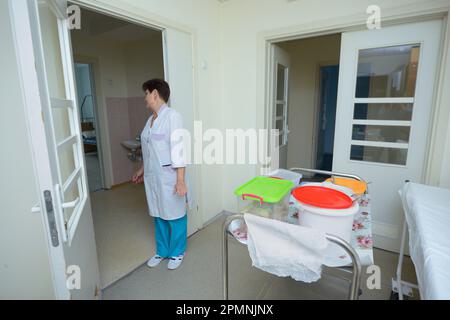 Infermiera in camici da laboratorio che tira un carrello a vassoio con cibo in pentole per i pazienti in una hall di un ospedale. Giugno 4, 2019. CityCenter di neurochirurgia. Kiev, U. Foto Stock