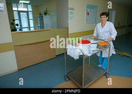 Infermiera in camici da laboratorio che tira un carrello a vassoio con cibo in pentole per i pazienti in una hall di un ospedale. Giugno 4, 2019. CityCenter di neurochirurgia. Kiev, U. Foto Stock