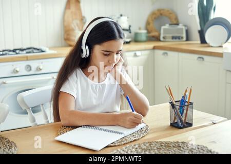 Graziosa ragazza pre-teen in cuffie wireless scrittura compito, rende compito sedersi a tavola in cucina, sembra focalizzato, impegnato in esercizio. Homeschooling Foto Stock