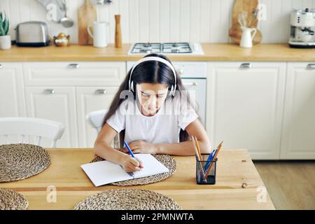 Graziosa ragazza pre-teen in cuffie wireless scrittura compito, rende compito sedersi a tavola in cucina, sembra focalizzato, impegnato in esercizio. Homeschooling Foto Stock