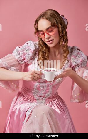 Ritratto di felice principessa affascinante indossando bel vestito e occhiali da sole tenendo una tazza di caffè e guardando su di esso su sfondo studio. Mattina Foto Stock