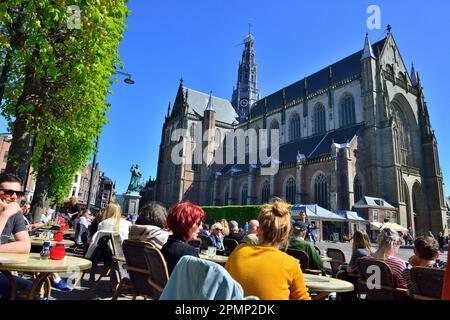 La gente gode del sole primaverile nei ristoranti esterni intorno alla piazza centrale del mercato (Grote Markt) oltre alla chiesa gotica Grote Kerk, Haarlem Foto Stock