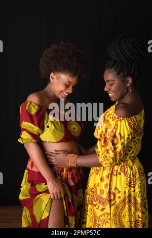 Due donne amiche, una delle quali incinta, indossando insieme abiti colorati. Isolato su sfondo giallo. Foto Stock