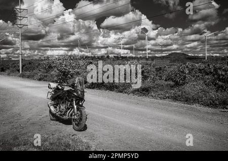 Una grande motocicletta nera parcheggiata su una pista sterrata accanto a una linea di trasmissione di energia, enormi turbine eoliche e una piantagione di caffè nella provincia di Gia Lai Foto Stock