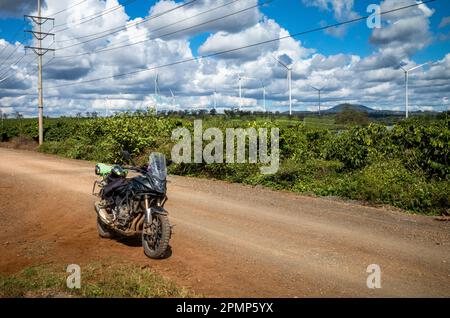 Una grande motocicletta nera parcheggiata su una pista sterrata accanto a una linea di trasmissione di energia, enormi turbine eoliche e una piantagione di caffè nella provincia di Gia Lai Foto Stock