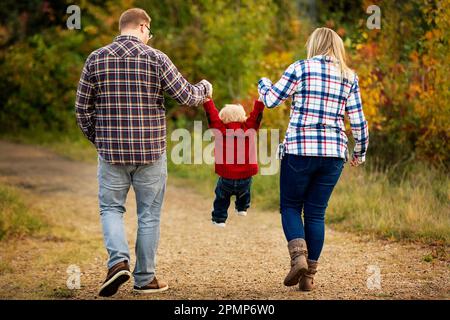 Madre e padre si tengono per mano con il figlio più piccolo mentre camminano lungo un sentiero in un parco cittadino in autunno; Edmonton, Alberta, Canada Foto Stock