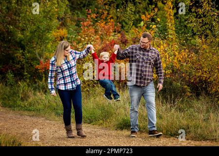 Madre e padre si tengono per mano con il figlio più piccolo mentre camminano lungo un sentiero in un parco cittadino in autunno; Edmonton, Alberta, Canada Foto Stock