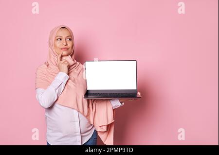 Donna musulmana incinta e penosa in hijab rosa, tiene il portatile con schermo bianco, ragionamento sul suo congedo di maternità Foto Stock