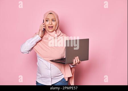 Donna musulmana araba incinta che lavora in hijab, parlando al telefono delle cellule, tenendo il laptop, telelavoro. Gravidanza e carriera Foto Stock