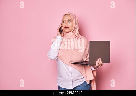 Elegante donna musulmana araba incinta in hijab rosa, negoziando con i partner commerciali sul telefono cellulare, tenendo il computer portatile Foto Stock
