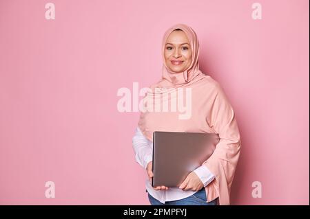 Donna musulmana araba fiduciosa in hijab rosa, con un notebook, sorridente guardando la fotocamera, sfondo rosa isolato Foto Stock