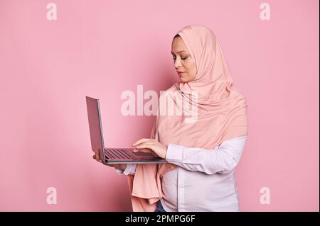 Multitasking arabo musulmano donna incinta in hijab rosa, digitando il testo sulla tastiera del computer portatile. Gravidanza, affari e carriera Foto Stock