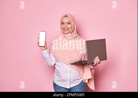 Donna musulmana araba incinta sorridente in hijab rosa, con smartphone con schermo bianco, sfondo rosa isolato Foto Stock