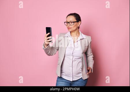 Donna ispanica in stato di gravidanza sicura che tiene in mano un notebook, controlla l'app mobile sul suo smartphone, sfondo rosa isolato Foto Stock