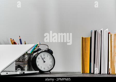 Concetto di ritorno a scuola o idea del giorno dell'insegnante. Penne, matite, libri, una sveglia e un computer portatile sul tavolo, sullo sfondo di una tavola grigia. Foto Stock