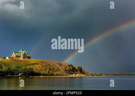 Doppio arcobaleno in un cielo scuro su un lago con uno storico edificio in legno sul fianco della collina che si affaccia sul lago, che si illumina di luce calda al tramonto Foto Stock