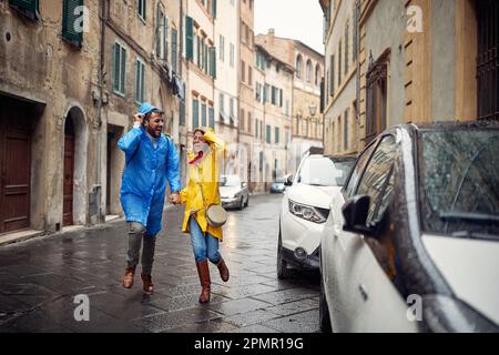 Una giovane coppia allegra in impermeabile sta scappare dalla pioggia mentre cammina per la città in un'atmosfera tranquilla. Passeggiata, pioggia, città, rapporto Foto Stock