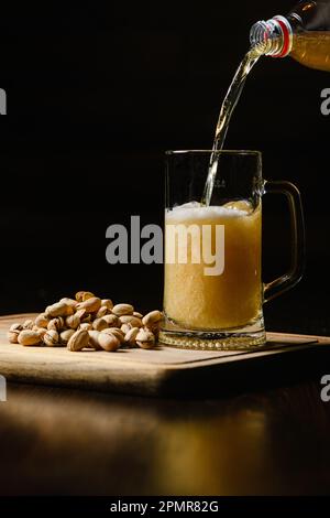 La birra viene versata in un bicchiere freddo su sfondo scuro. Primo piano. Una manciata di pistacchi si trova accanto alla tazza. Foto Stock