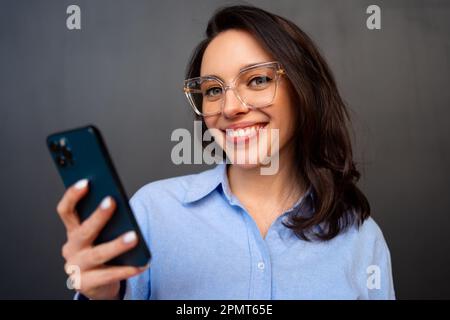 Ritratto sorridente donna che tiene in mano lo smartphone guardando la fotocamera, indossare occhiali, digitare messaggi di testo sul telefono cellulare, gode di comunicazione online, su sfondo nero studio. Foto Stock